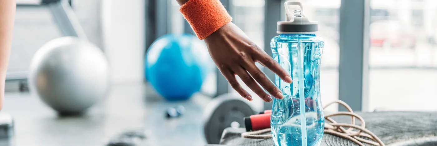 A gym floor with dumbbells and exercise balls, someone reaching for a sports water bottle