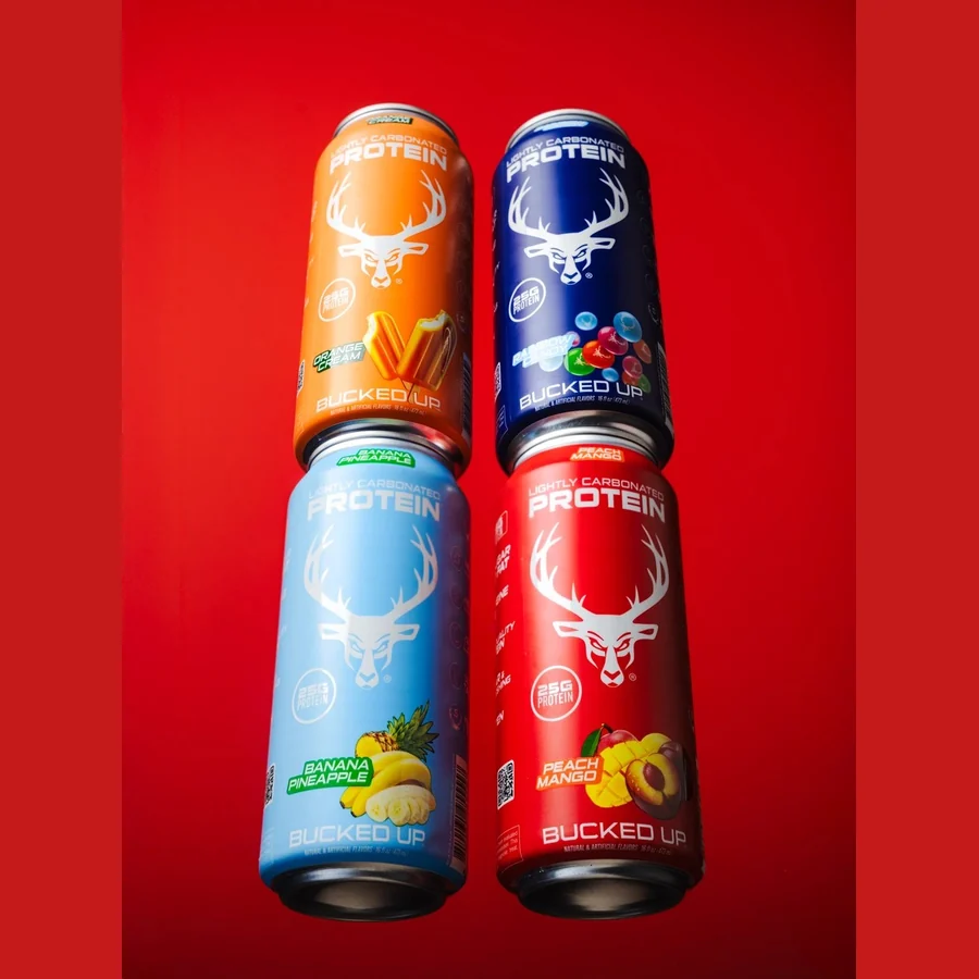 Four Bucked Up Protein Soda cans stacked on a red background, showing the Banana Pineapple, Berry, Blue Raspberry and Peach Mango labels
