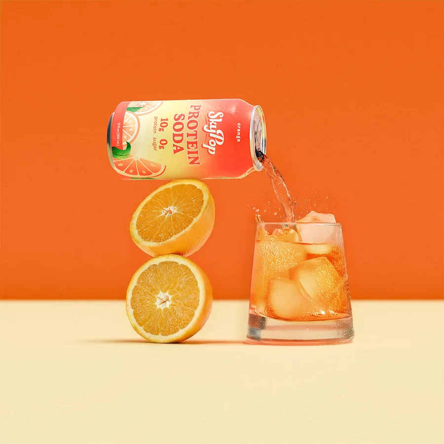 SkyPop Orange protein soda can standing among fresh orange slices beside a glass of orange soda over ice